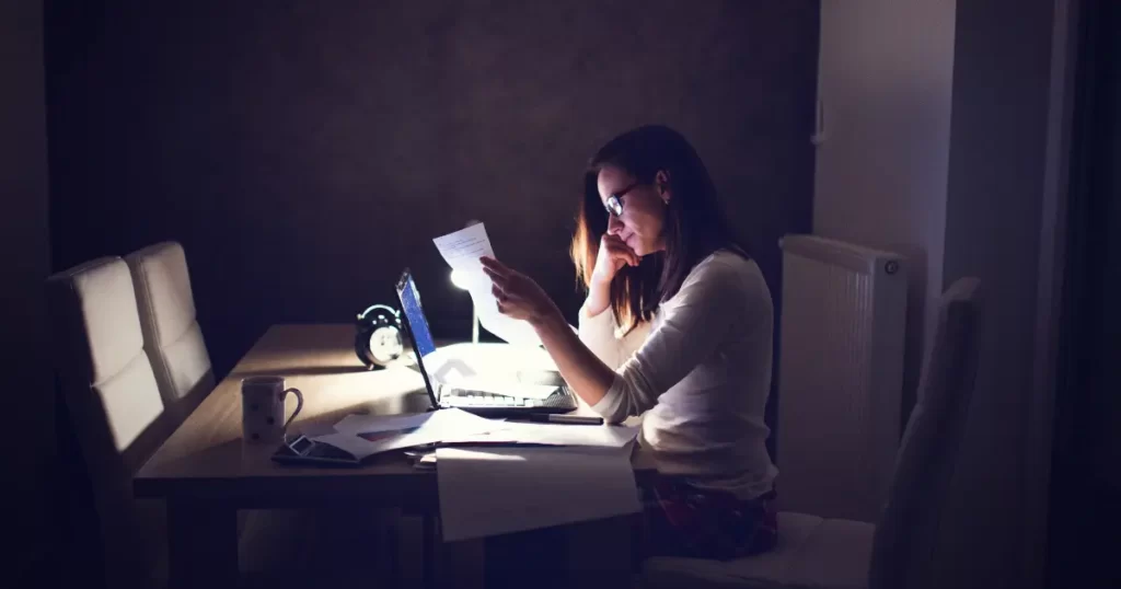 Working woman at desk in dark room displaying masculine (wounded) energy (workaholic)